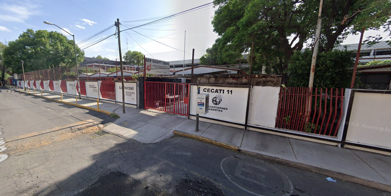 Fotografía en la que se muestra la entrada del plantel, conformada por muros de color blanco intercalados con rejas de color rojo. Una de las rejas es la entrada al plantel y debajo de ella hay una rampa de acceso. En uno de los muros blancos, en letras grandes negras dice: CECATI 11.
